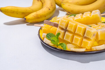 Bright yellow vegan banana ice cream popsicles. Frozen banana fruit lollypops, with ice and mint decor, white kitchen background. Summer cold vegetarian healthy dessert, copy space