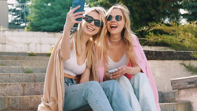 Two young beautiful blond smiling hipster women in trendy summer clothes. Carefree models posing on street background. Positive models having fun. Holding smartphone, take selfie photos