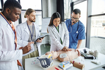 young asian woman in white coat doing chest compressions on CPR manikin near instructor, medical equipment and multicultural students with notebooks, emergency situations response concept