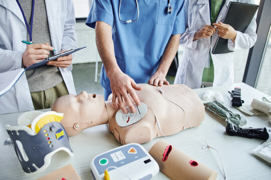 cropped view of instructor applying defibrillator pads on CPR manikin near medical equipment and young students in white coats during first aid seminar, life-saving skills hands-on learning concept