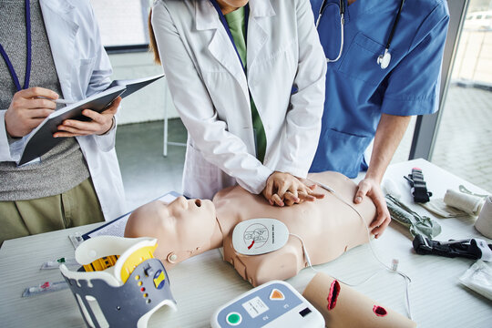 first aid seminar, partial view of young woman in white coat practicing chest compressions on CPR manikin near paramedic and student writing in notebook, life-saving skills hands-on learning concept