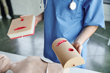 partial view of medical instructor showing wound care simulators while standing near CPR manikin in training room, first aid hands-on learning and critical skills development concept
