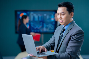Handsome business man and suit holding the laptop in his hands and writing something. Side view, standing confident in the office in front of his team.