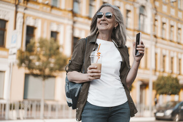 Obraz premium concept of travel in old age, featuring mature woman enjoying trip in old city of Europe. senior woman seen appreciating view of city and capturing moment by taking photos on mobile phone.