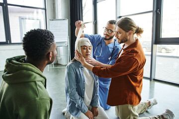 Obraz premium young man and medical instructor bandaging head of asian woman near african american participant of first aid seminar in training room, importance of emergency preparedness concept