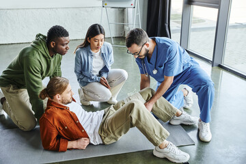 african american man and asian woman looking at medical instructor applying compressive bandage on leg of participant of first aid training seminar, bleeding prevention techniques concept