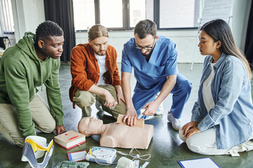 healthcare worker tamponing wound on simulator with bandage while showing life-saving skills to multicultural team near CPR manikin, defibrillator and medical equipment, emergency response concept