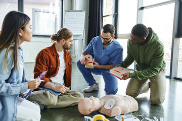 medical instructor and african american man holding wound care simulators near CPR manikin, defibrillator and multiethnic participants with notebook and clipboard, life-saving skills concept - obrazy, fototapety, plakaty