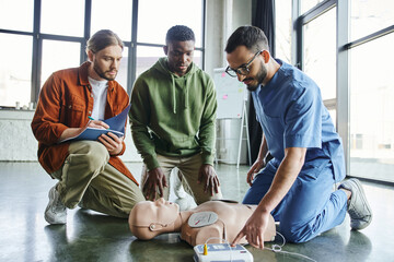 cardiac resuscitation, young man writing in notebook near african american participant and medical instructor operating defibrillator on CPR manikin during first aid training seminar - obrazy, fototapety, plakaty