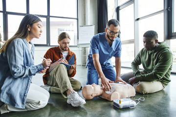cardiac resuscitation skills, healthcare worker applying defibrillator pads on CPR manikin near young diverse and multiethnic group, health care and life-saving techniques concept