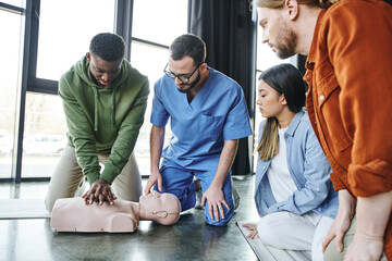 young multiethnic people and medical instructor looking at african american man doing chest compressions on CPR manikin, cardiopulmonary resuscitation, first aid training seminar
