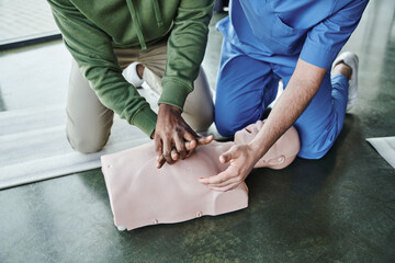 cropped view of african american man practicing life-saving skills and doing chest compressions on CPR manikin near professional paramedic on first aid seminar, emergency preparedness concept