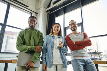 young and successful multiethnic business colleagues smiling at camera in modern office, african american man with laptop, asian woman with takeaway drink, bearded man in eyeglasses