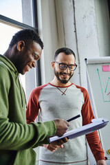 young and cheerful african american entrepreneur showing documents on clipboard to african american business partner in eyeglasses while working on startup in office, successful collaboration