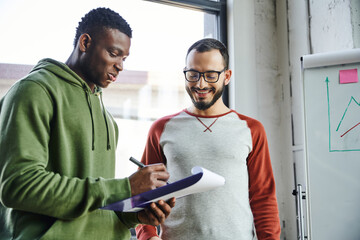 young african american businessman in green hoodie writing on clipboard near smiling bearded colleague in eyeglasses and flip chart with graphs in modern office, creative youth © LIGHTFIELD STUDIOS