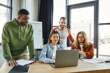 happy asian businesswoman working on laptop near amazed multiethnic men, documents and flip chart with graphs on background in modern office, creative team collaboration