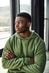 pensive african american businessman in green hoodie standing with folded arms and looking away near window in modern office, young entrepreneur