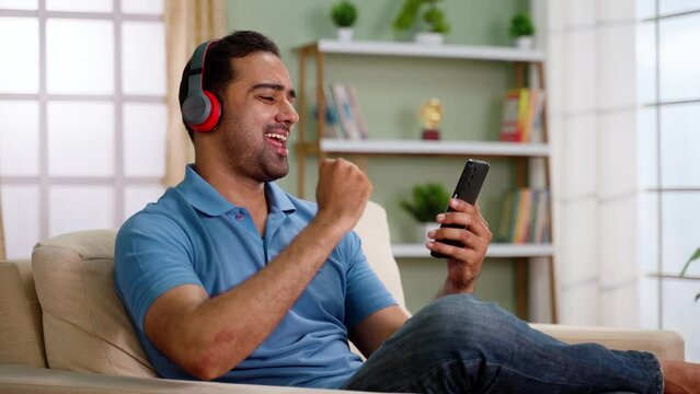 Cheerful indian man with wireless headphones listening favorite songs on mobile phone on sofa at home - concept of Music enthusiast, joyful experience and relaxation