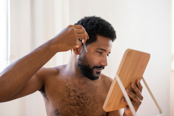 Young bearded African American young man looking at himself in a small wooden mirror, and applying...