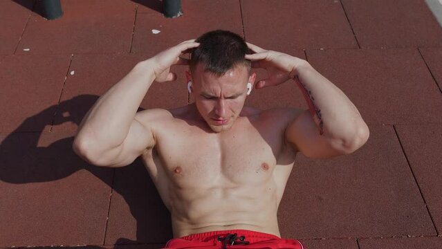 Slow motion handheld shot from above of fit shirtless sportsman with earbuds lying on ground doing crossed arms crunch exercise during outdoor training session