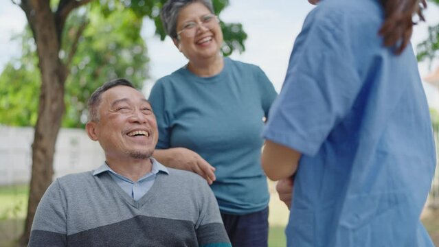 Happy asian adult male patient enjoy talking with his wife and woman caregiver nurse while sitting on wheelchair relaxing at hospital park. Retirement, physiotherapy and health care