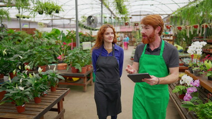 Two Staff colleagues walking through flower shop holding tablet. Happy redhaired man and woman wearing green apron inside plant local business store