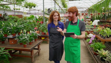 Fototapeta na wymiar Two Staff colleagues walking through flower shop holding tablet. Happy redhaired man and woman wearing green apron inside plant local business store
