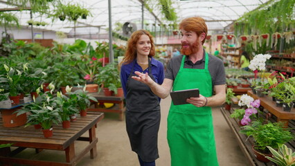 Two Staff colleagues walking through flower shop holding tablet. Happy redhaired man and woman wearing green apron inside plant local business store