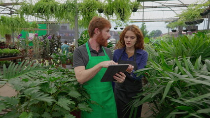 Two entrepreneurs talking about plant business while walking through isles wearing aprons. A male employee explaining Flower Shop procedure to female colleague while holding tablet