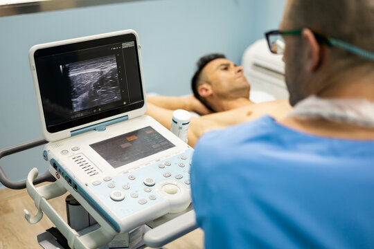 A physical therapist in a blue uniform and eyeglasses performs an ultrasound. The patient's face is seen in the background. Ultrasound concept in injured athletes Modern ultrasound in medical center. - Powered by Adobe