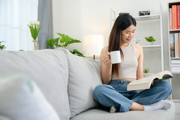 Obraz premium Asian woman at home sitting and reading a book on the sofa in the living room Smiling young woman relaxing in her living room reading a book and drinking coffee.