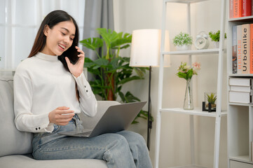 Asian woman talking on the phone happy looking at laptop sitting on the sofa in the living room at home and work online with a laptop computer feeling excited and happy