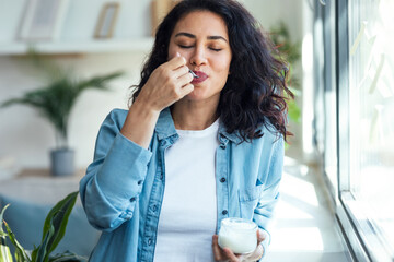 Happy beautiful woman eating yogurt while standing in living room at home.