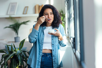 Happy beautiful woman eating yogurt while standing in living room at home.