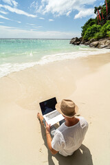 Man in the summer hat , a businessman, digital nomad working with laptop on the sandy beach. Freelancer. Tropics.