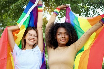 Happy smiling homosexual lesbian couple holding rainbow flag, beautiful woman and her African curly hair girlfriend celebrating pride month together, romantic LGBT lover and gay pride movement concept