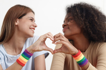 Hand making a heart sign wearing LGBT rainbow flag wristband, happy smiling homosexual lesbian couple looking at each other as blurred background, romantic LGBT lover celebrating pride month together.