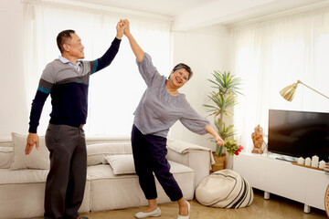 Obraz premium Happy smiling Asian elderly couple dancing in living room, romantic senior lover having fun together at home, mature retired grandfather grandmother celebrate wedding Anniversary, retirement lifestyle