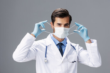 A male doctor in a white coat and medical mask and sterile gloves looks at the camera on a gray isolated background, copy space, space for text