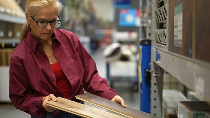 Attractive mature woman shopping for wood flooring and compares colors in a hardware store shopping...