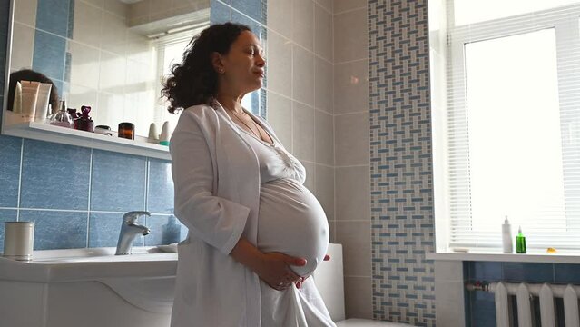 African American adult pregnant woman in white bathrobe, standing in the home bathroom, smiling looking aside and gently stroking her belly, enjoying happy moments of her pregnancy and maternity