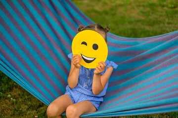 Obraz na płótnie Canvas The girl hides face behind a round yellow cheerful winking emoticon while resting on a hammock on a warm summer day. A charge of good mood and jovial spirit