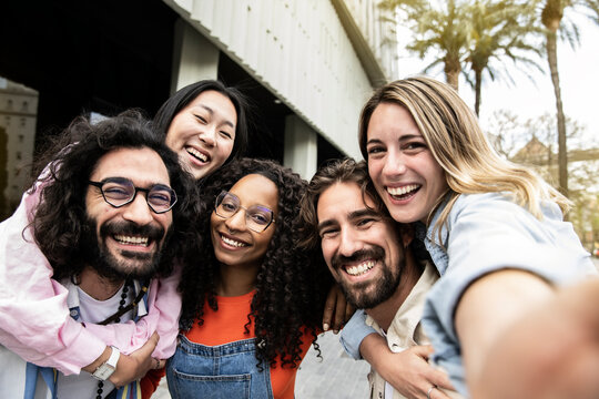 Diverse group of friends laughing hugging each other and taking a selfie in the street.Cheerful multiracial group of young hipsters taking a picture outside and looking at camera.