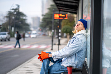 Tired guy tourist with coffee take away waiting bus. Blonde hair millennial man on bus stop in...