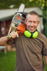Middle-aged gardener holding a chainsaw over his shoulder after work with ear protection looking at the camera