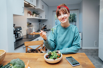 Smiling young with woman with opened daily veganuary calendar on phone app while eating fresh green...
