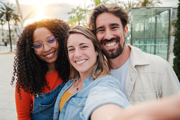 Three young adult friends smiling taking a selfie portrait and having fun together. Group of multiracial cheerful people celebrating their friendship. Two women and one man with positive expression - obrazy, fototapety, plakaty