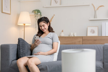 Happy pregnant asian woman use mobile phone to watch video media and relax with social media online.Pregnant woman smile and laughing lying on couch resting at home.Pregnancy and Technology Concept