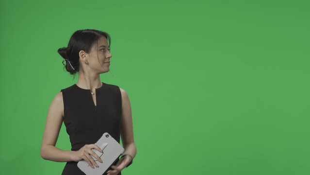 TV presenter against a green screen. Asian Anchorwoman with a tablet in his hands speaks, points to the green screen, listens, nods. Advertising area, workspace mock up. HDR BT2020 HLG Material.