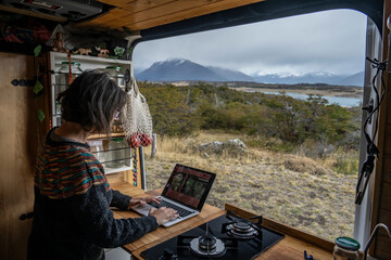 Elderly woman works on her laptop inside a motorhome in front of a scenic landscape in the area of Lake Roca, Santa Cruz, Argentine Patagonia.